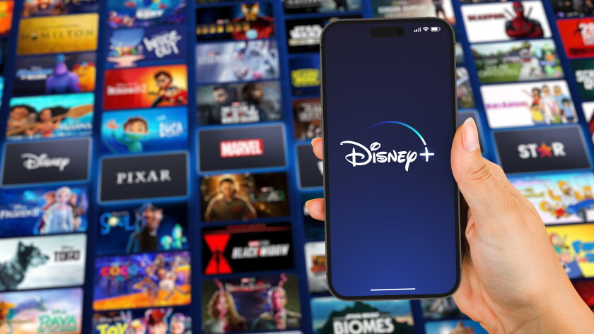 Female hand holding a smartphone iPhone 14 Pro with Disney Plus (Disney+) streaming app app on the screen. TV blurred on background. Rio de Janeiro, RJ, Brazil. September 2022